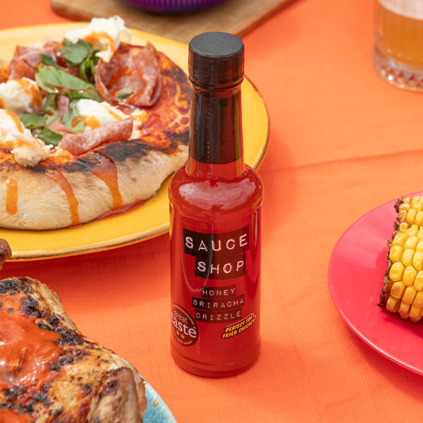 A bottle of Sauce Shop Honey Sriracha Drizzle surrounded by mozzarella pizza, corn on the cob and chicken 