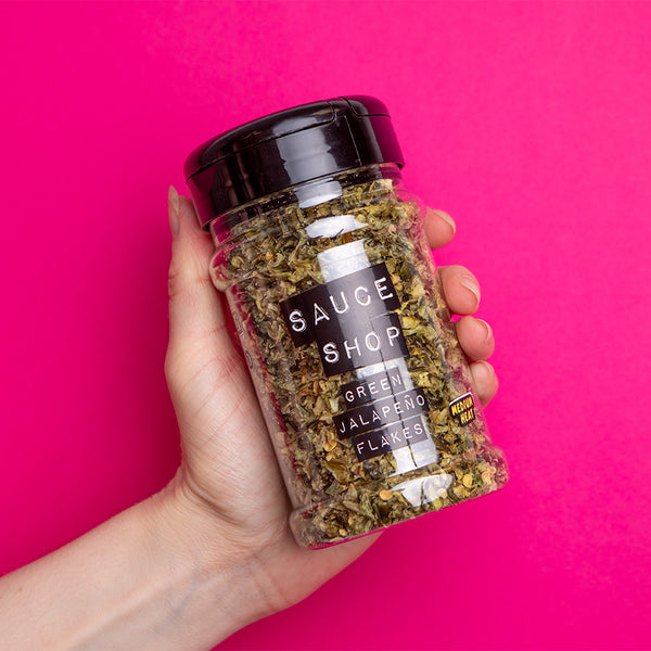 A shaker jar of Sauce Shop Green Jalapeno Flakes being held against a pink background