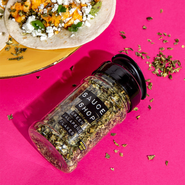 A shaker jar of Sauce Shop Green Jalapeno Flakes laying on a pink table next to spicy tacos