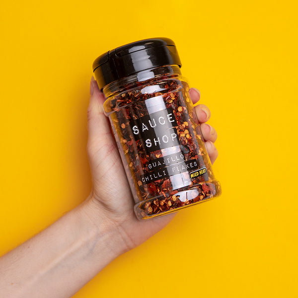 A shaker jar of Sauce Shop Guajillo Chilli Flakes being held up against a yellow background