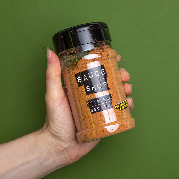 A shaker jar of Sauce Shop Original BBQ Rub being held by a hand in front of a green background