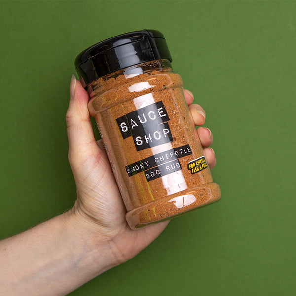 A shaker jar of Sauce Shop Smoky Chipotle BBQ Rub being held by a hand in front of a green background