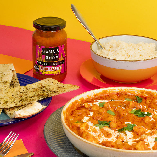 A jar of Sauce Shop Special Makhani Simmer Sauce with a bowl of rice, chickpea curry and naan bread on a pink and yellow background